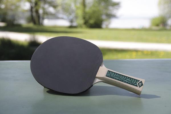 Tischtennisschläger mit schwarzem Belag auf grünem Tisch im Freien bei Sonnenschein