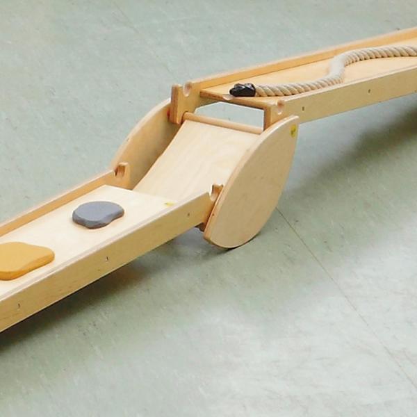 Holzrutsche mit Seil und farbigen Steinen auf grünem Boden für motorische Übungen.