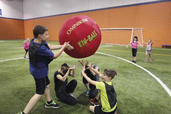Kinder spielen Kin-Ball in der Halle auf Kunstrasen mit großem roten Ball.
