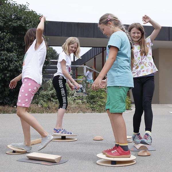 Vier Mädchen balancieren auf Holz-Balance-Boards im Freien vor einem Gebäude.