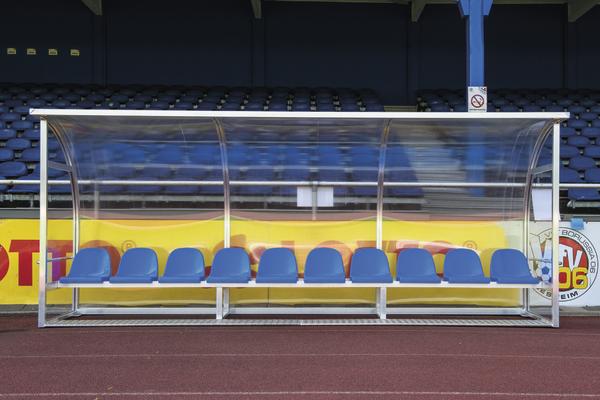 Leere Spielerbank mit blauen Sitzen und transparentem Wetterschutz im Stadion.