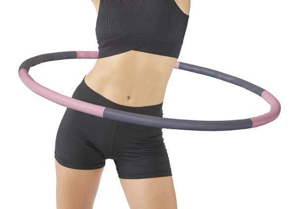 Frau trainiert mit pink-grauem Hula-Hoop-Reifen in schwarzer Sportkleidung vor weißem Hintergrund.