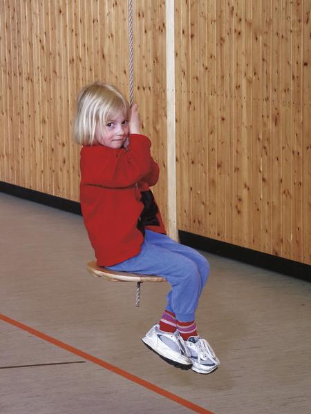 Kind schaukelt an Seil in Turnhalle mit Holzverkleidung, trägt rote Jacke und blaue Hose.