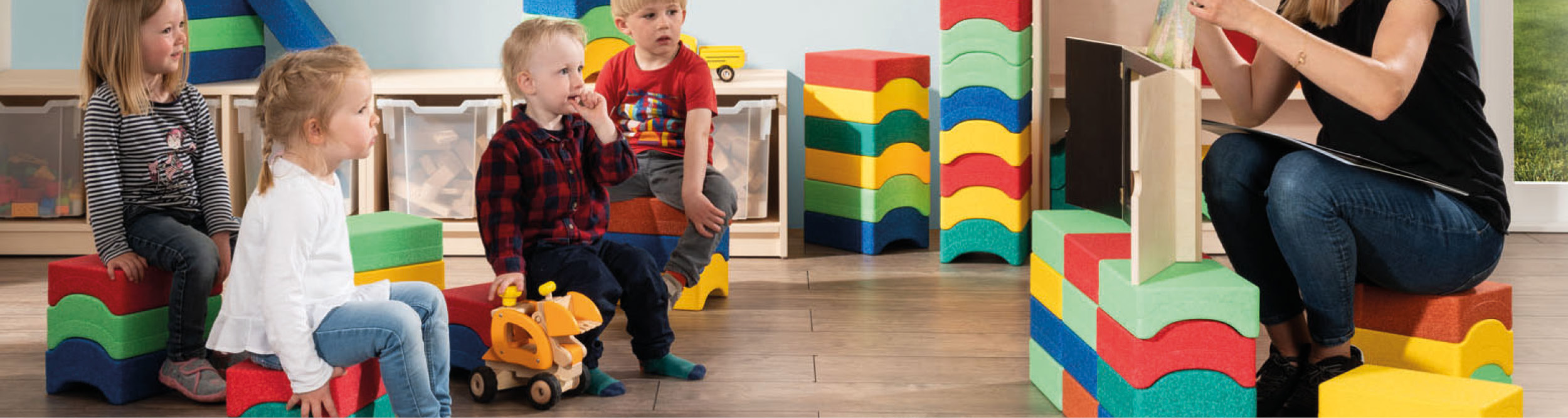 Erzieherin zeigt einer kleinen Kindergruppe auf bunten Schaumstoffhockern etwas auf einem Blatt Papier.