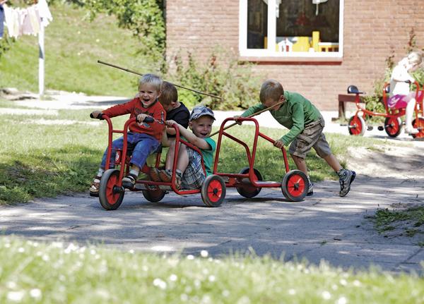 Kinder spielen draußen mit roten Tretfahrzeugen auf einem sonnigen Hof vor einem Backsteinhaus.