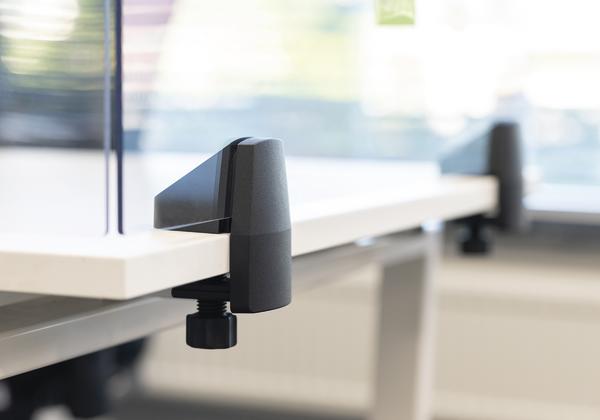 Trennwand aus Glas mit schwarzer Klemme auf weißem Bürotisch, Bürotrennung.