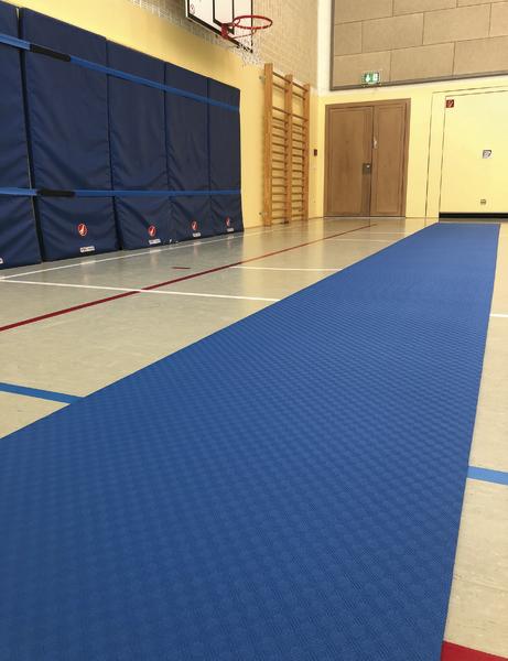 Blauer Turnmatten-Laufweg in einer Sporthalle mit Basketballkorb und Wandpolstern.