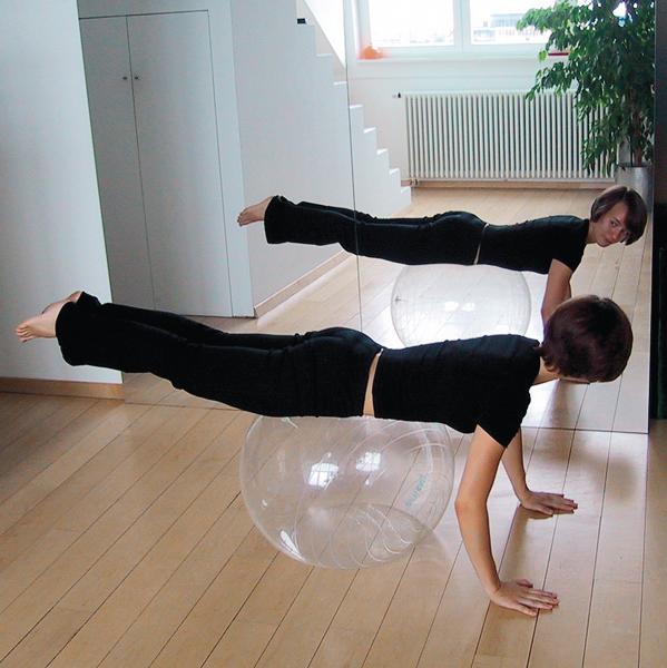 Frau macht Rückenübung auf Gymnastikball vor Spiegel in hellem Raum mit Holzfußboden.