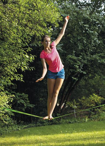 Frau balanciert auf Slackline im Garten, umgeben von grünen Bäumen und Gras.