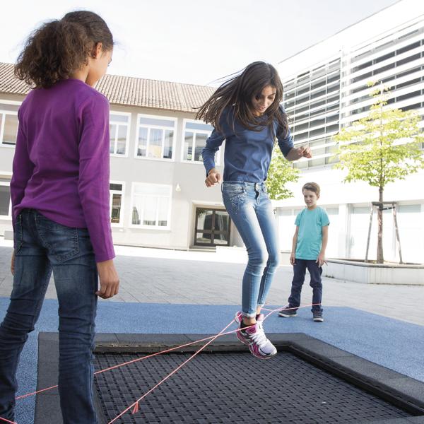 Kind spielt auf Trampolin mit gespannten Gummibändern, andere Kinder beobachten draußen.