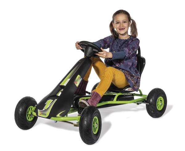 Mädchen fährt lachend grünes Pedal-Kart mit schwarzem Lenkrad auf weißem Hintergrund.