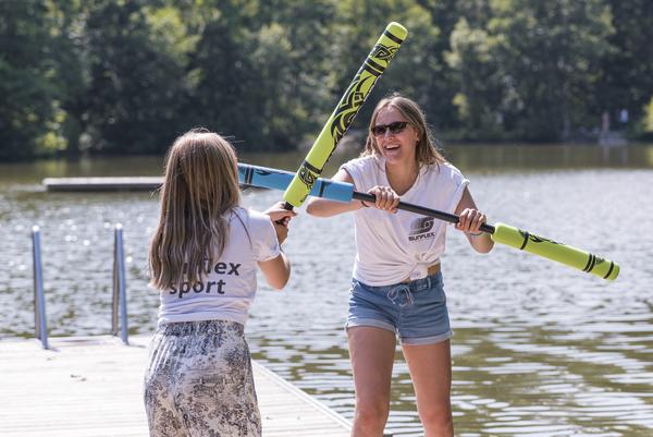Zwei Mädchen kämpfen mit Schaumstoffschwertern auf einem Steg am See bei Sonnenschein.