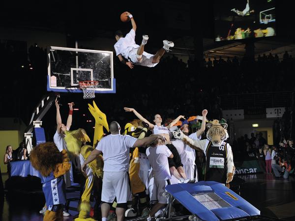 Basketballspieler springt für Dunking über mehrere Maskottchen bei Show-Event in Halle.