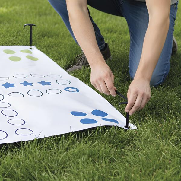 Person befestigt die Ecke einer Spielmatte im Gras mit einem Erdanker.