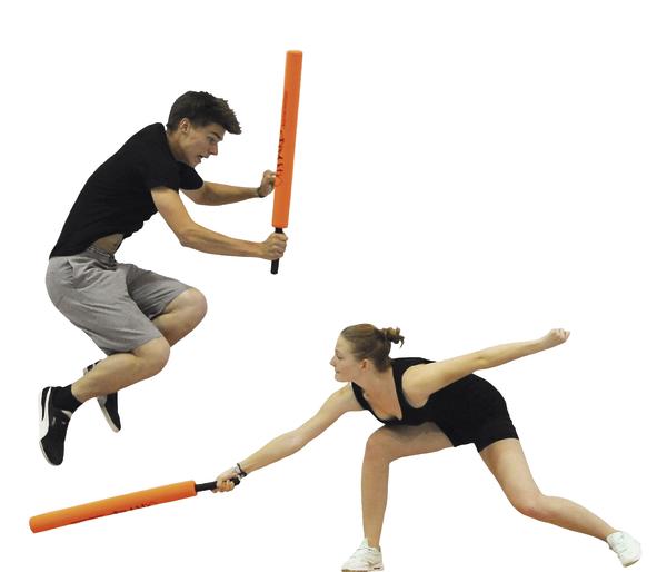 Zwei Personen kämpfen dynamisch mit Schaumstoffstäben, eine springt, die andere weicht aus.