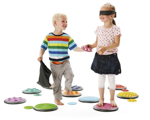 Zwei Kinder spielen barfuß mit bunten Balancierplatten, ein Mädchen trägt eine Augenbinde.