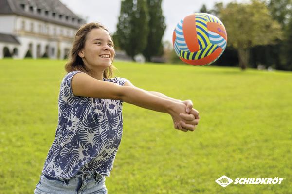 Mädchen spielt Volleyball im Park, schlägt bunten Ball mit verschränkten Armen.