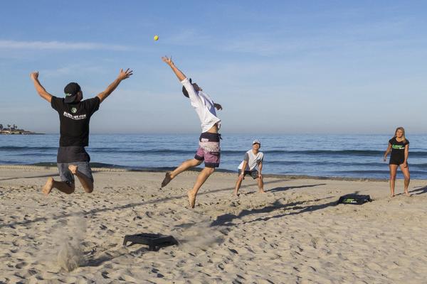Vier Personen spielen Beach-Tennis am Strand bei klarem Himmel und ruhigem Meer.