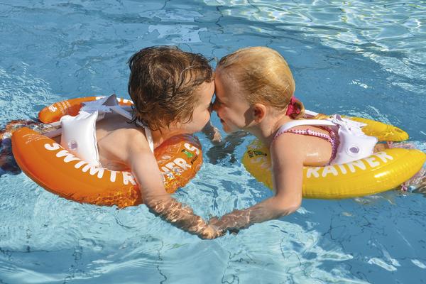 Zwei Kinder mit Schwimmringen halten sich im Pool an den Händen und berühren sich mit der Stirn.