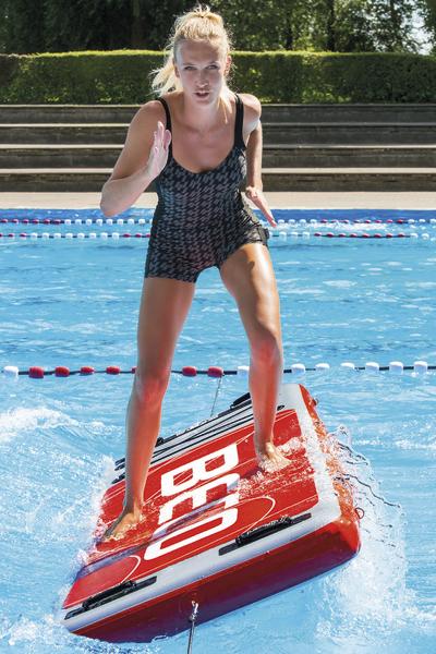 Frau balanciert auf rotem Schwimmbrett im Pool, trägt schwarzen Badeanzug.