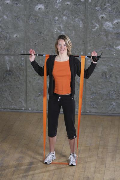 Frau trainiert mit Widerstandsband und Stab vor grauer Wand in Sportkleidung.