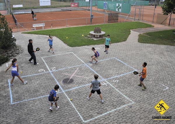 Kinder spielen Street Racket auf einem markierten Spielfeld im Freien neben Tennisplätzen.