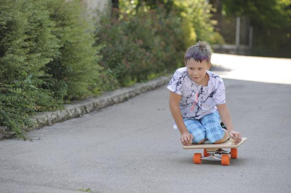 Junge fährt auf einem Longboard sitzend auf einem Gehweg in einer grünen Wohngegend.