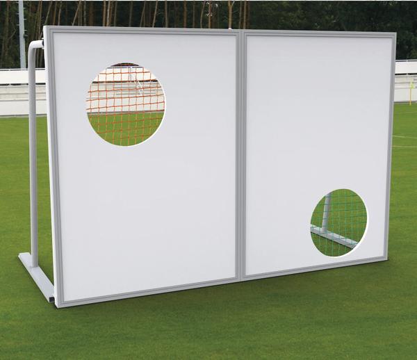 Torwand mit zwei unterschiedlich großen Löchern auf grünem Fußballfeld vor Waldkulisse.