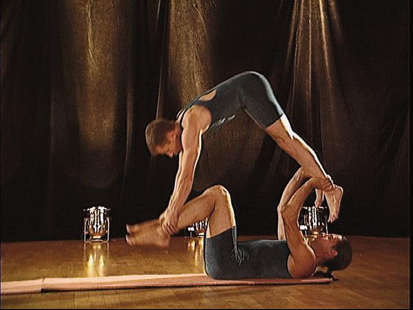 Zwei Männer führen Partnerakrobatik auf Matte in Studio mit dunklem Vorhang aus.