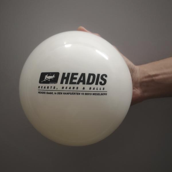 Hand hält weißen HEADIS Ball mit schwarzem Logo vor grauem Hintergrund.