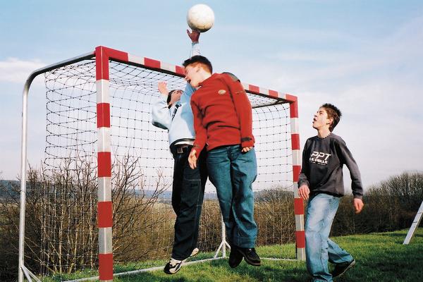 Drei Jungen spielen Handball, einer springt und hält den Ball über dem Tor.