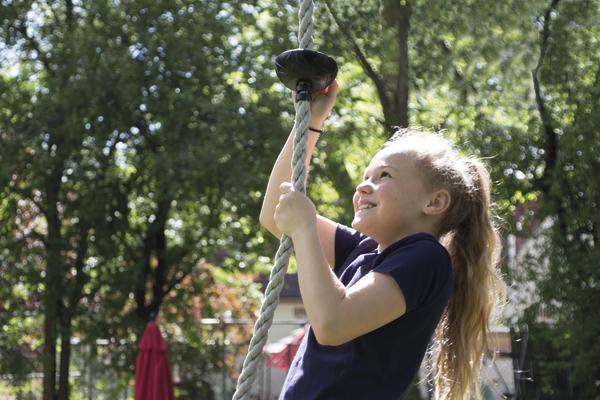 Mädchen klettert fröhlich an einem Seil im sonnigen Garten mit grünen Bäumen.