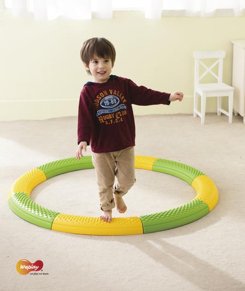 Kind balanciert barfuß auf grün-gelbem Balancierkreis im hellen Raum mit Stuhl.