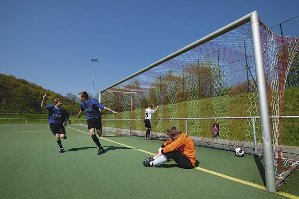 Junge Fußballspieler feiern Tor, während Torwart enttäuscht auf dem Spielfeld sitzt.