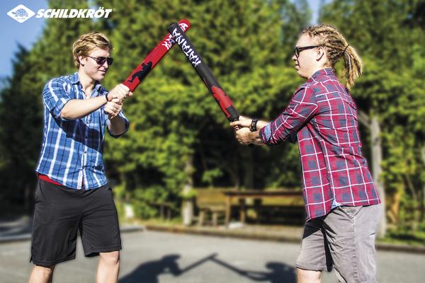 Zwei junge Männer kämpfen mit Schaumstoffschwertern im Freien bei Sonnenschein.