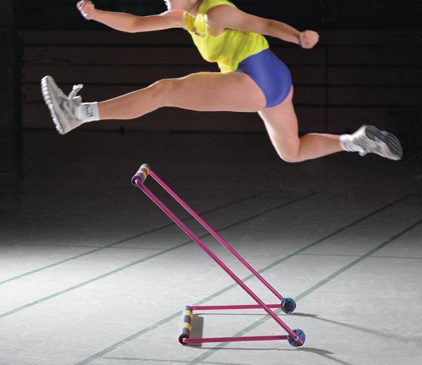 Frau in Sportkleidung springt über pinken Hürdenständer in Sporthalle.