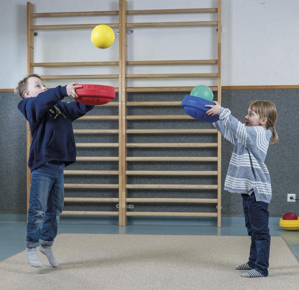 Zwei Kinder werfen in einer Turnhalle Bälle mit bunten Schaumstoffscheiben in die Luft.