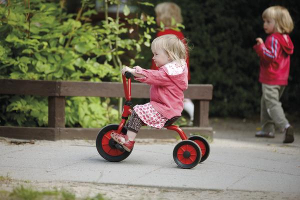 Kind auf rotem Dreirad im Park, zwei weitere Kinder im Hintergrund spielend.
