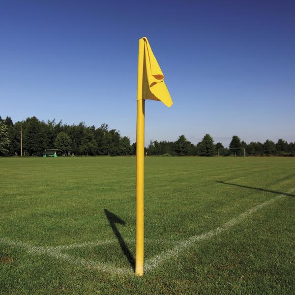 Gelbe Eckfahne auf Fußballplatz, grüne Rasenfläche und blauer Himmel.