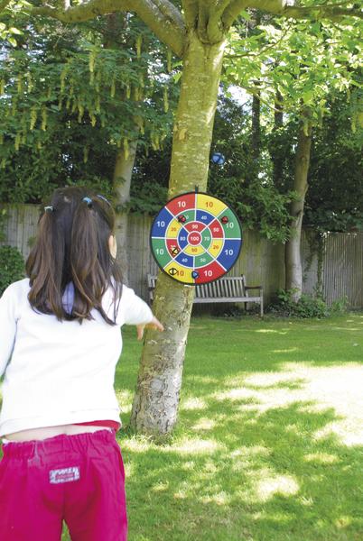 Kind wirft auf Dartscheibe am Baum im sonnigen Garten mit grüner Wiese und Bänken.