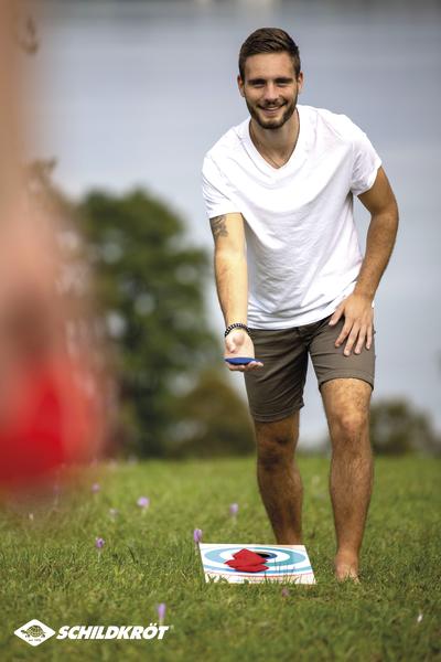 Junger Mann spielt Dosenwerfen im Freien auf einer Wiese bei Sonnenschein.