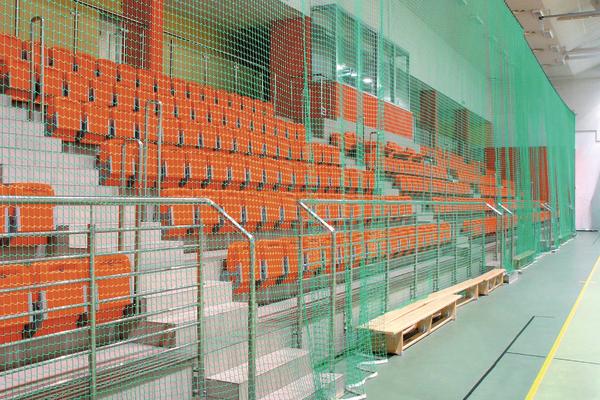 Leere orange Sitzreihen in einer Sporthalle hinter grünem Schutznetz.
