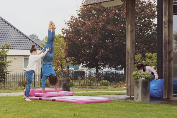 Zwei Mädchen üben Handstand auf Gymnastikmatten im Garten, eine hilft, die andere rollt Matte.