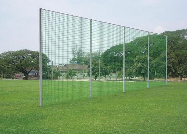 Hohe grüne Schutznetzwand auf Rasenfläche im Freien vor Bäumen und Gebäuden.
