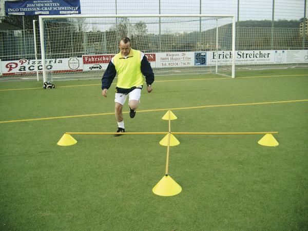 Mann trainiert Koordination mit Hürden und Kegeln auf Fußballplatz vor Tor.