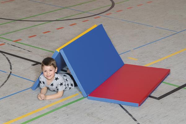Kind liegt lächelnd unter buntem Turnmatten-Tunnel in Sporthalle mit Linienboden.
