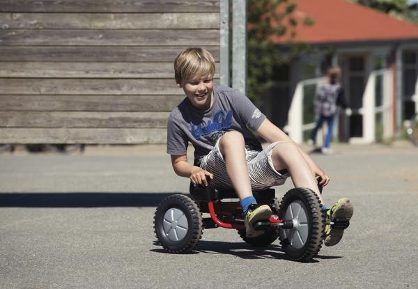 Junge fährt lachend auf einem roten Tretfahrzeug auf Asphalt im Freien.