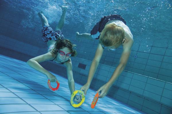 Kinder tauchen im Schwimmbad und spielen mit bunten Tauchringen unter Wasser.