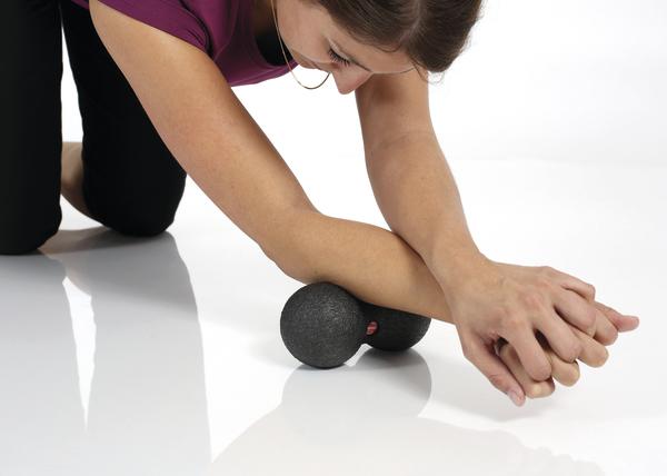 Frau kniet am Boden und nutzt einen Doppelball zur gezielten Selbstmassage des Unterarms zur Lockerung der Muskulatur.