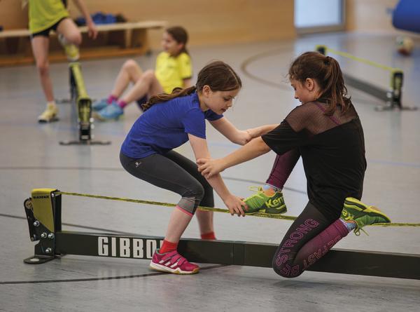 Kinder üben Slacklining in der Turnhalle, zwei Mädchen balancieren gemeinsam auf dem Gibbon-Band.